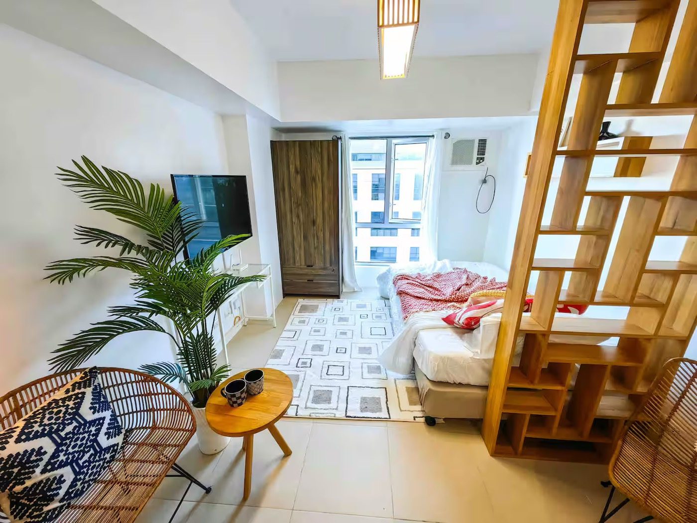 A finished Siempre Stays studio: a bed beside a full-height window, a timber room divider, rattan seating and a patterned rug.