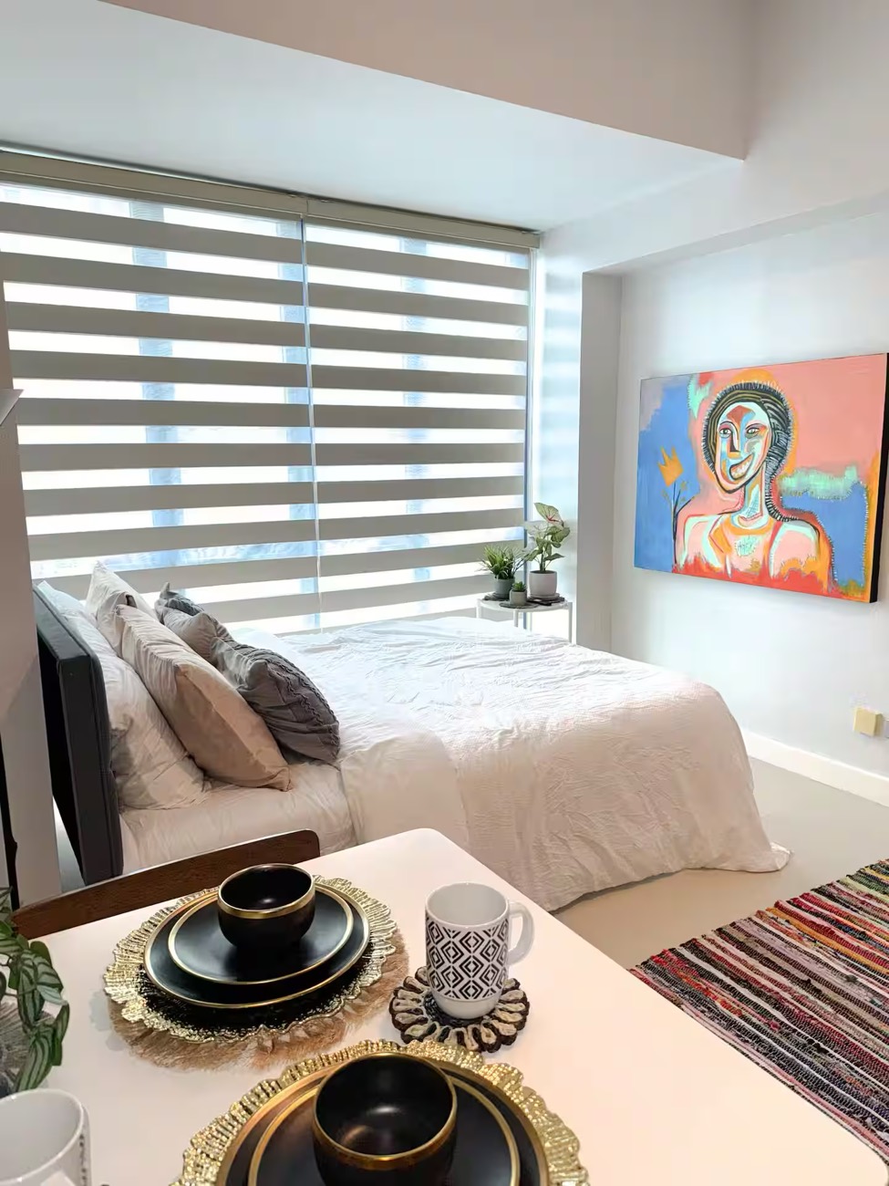 Estudio Basca: a made bed against Korean blinds, with a large expressionist portrait on the wall.