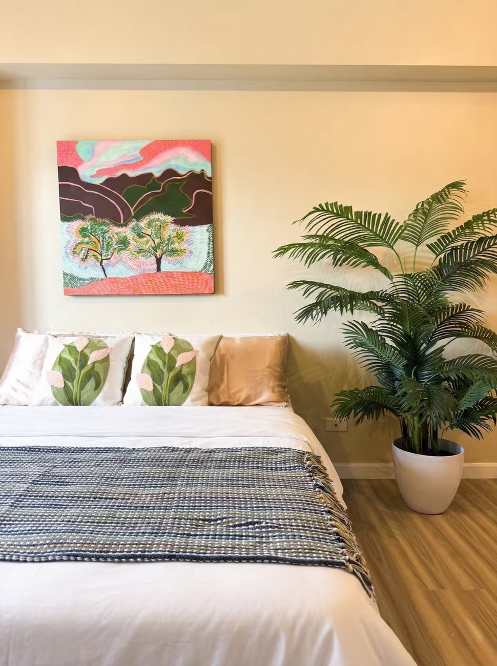 Estudio Serra: a bed dressed in white and a woven throw, beneath a framed mountain landscape, beside a tall palm.