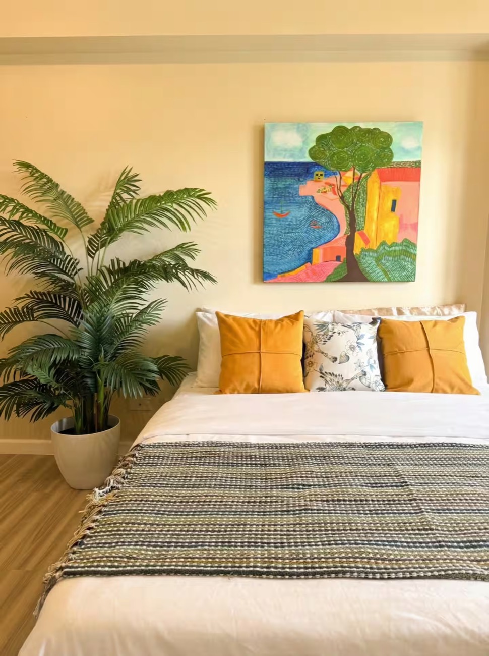 Studio Araya: a bed dressed with mustard cushions beneath a framed painting, next to a tall palm.