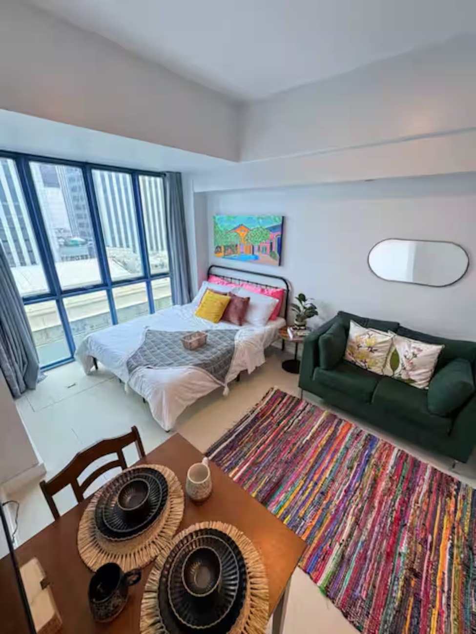 Vida Verde Studio: a bed beside floor-to-ceiling windows, a green velvet sofa, a colourful woven rug and a dining table set for two.