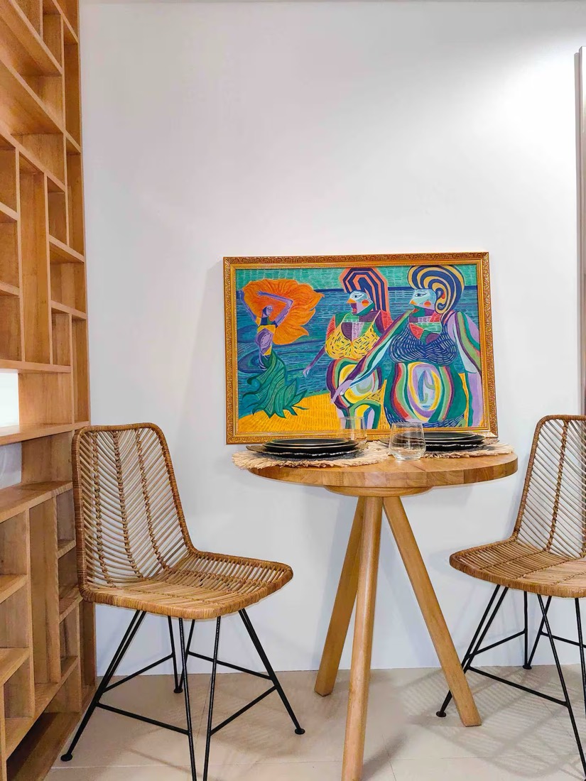 A framed painting above a round timber table set with dark ceramics and rattan chairs, in a Siempre Stays studio.